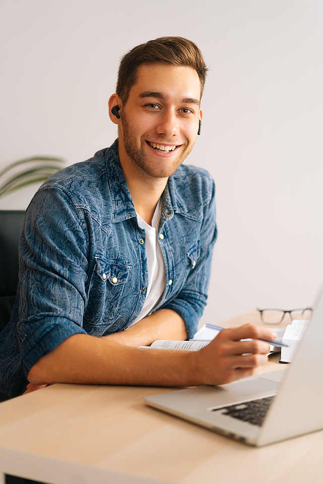Vertical shot of cheerful young business man in earphone having online video conference on laptop at home office.